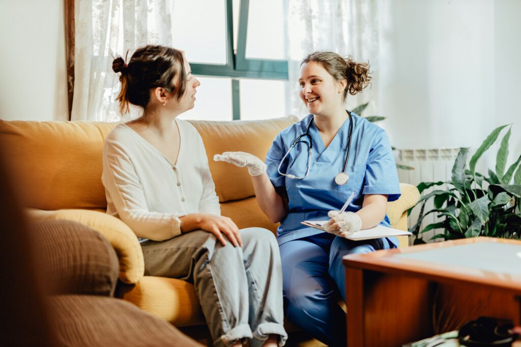 Healthcare worker in blue scrubs and a stethoscope sitting beside a woman on a couch during an in-home care visit, smiling and holding a clipboard while having a friendly conversation in a bright living room.