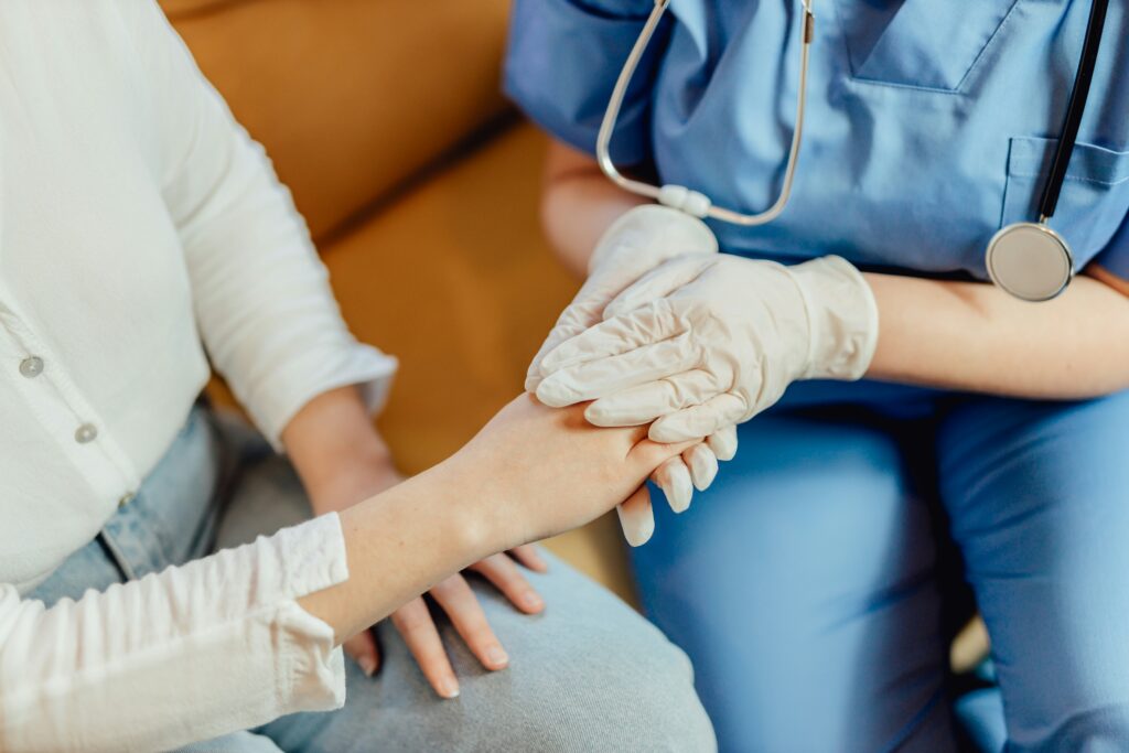 Close-up of a healthcare worker in blue scrubs and gloves gently holding a patient’s hand during an in-home care visit, with a stethoscope visible in a warm and supportive setting.