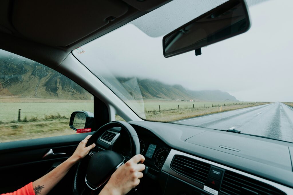 View from inside a car driving along a long rural road on an overcast day, with the driver’s hands on the steering wheel and misty mountains visible through the windshield in the distance. The image captures the dashboard, rearview mirror, and side window, creating a calm road trip atmosphere with open landscapes and cloudy weather.