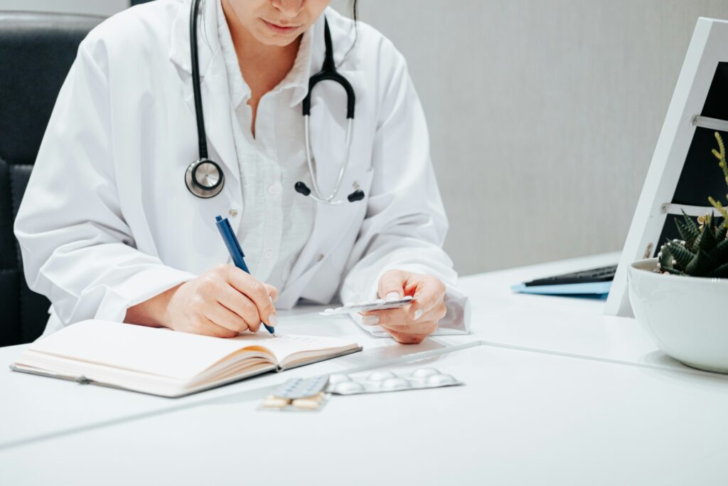 Doctor in a white coat writing notes at a desk while holding a blister pack of medication, with medical tablets and a stethoscope visible in a clinical office setting.