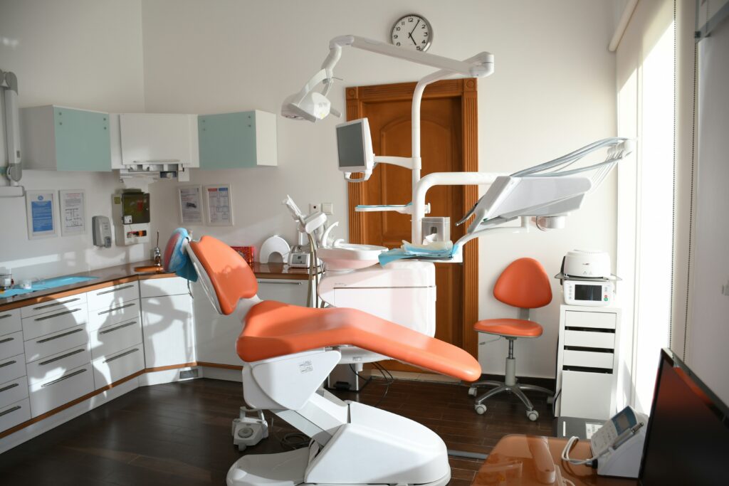 Modern dental clinic with an orange treatment chair, overhead examination lights, dental equipment, and clean cabinetry in a bright treatment room.