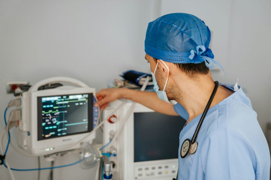 A medical professional wearing surgical scrubs, a blue cap and face mask monitors a patient’s vital signs on a bedside screen in a clinical environment.