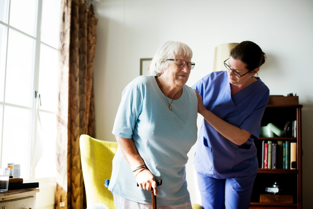 Healthcare worker in blue scrubs assisting an elderly woman with a walking cane in a sunlit living room, providing support as she stands beside a chair.