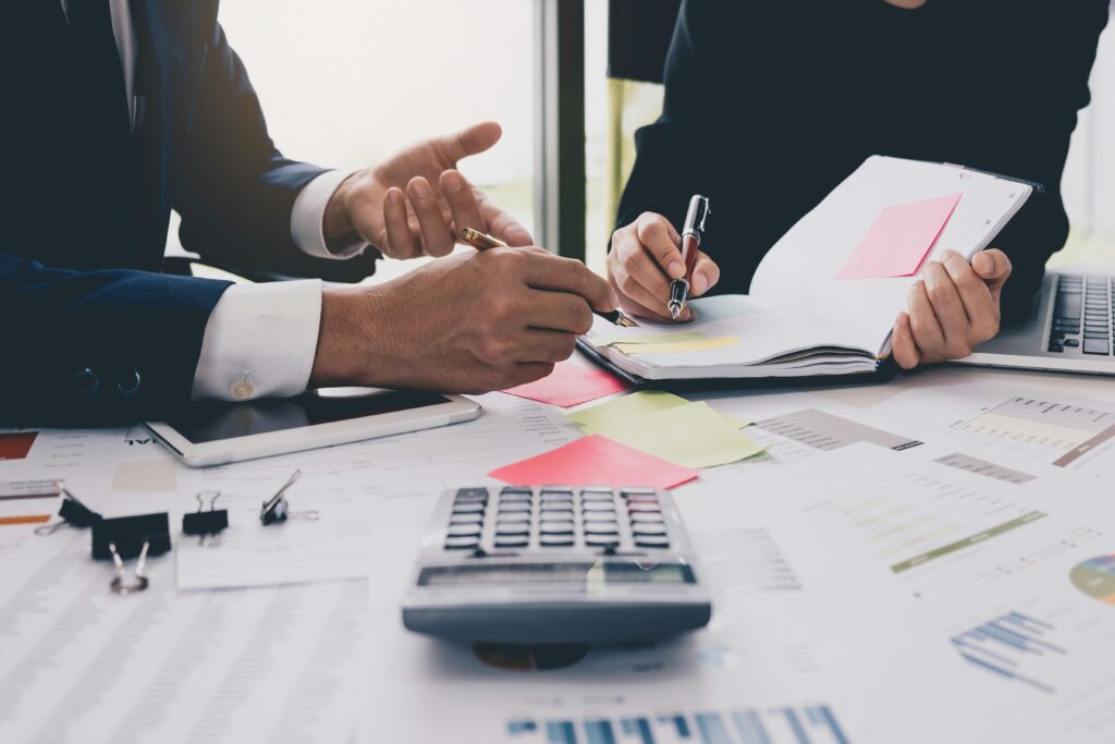 Professional office meeting with two people reviewing financial documents at a desk covered in paperwork, charts, sticky notes, and a calculator. One person gestures while discussing figures as the other takes notes in a notebook beside a laptop and tablet.