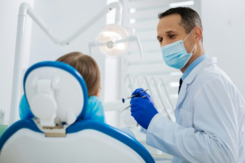 Dentist wearing a mask and gloves speaking with a patient in a dental chair while holding dental examination tools.