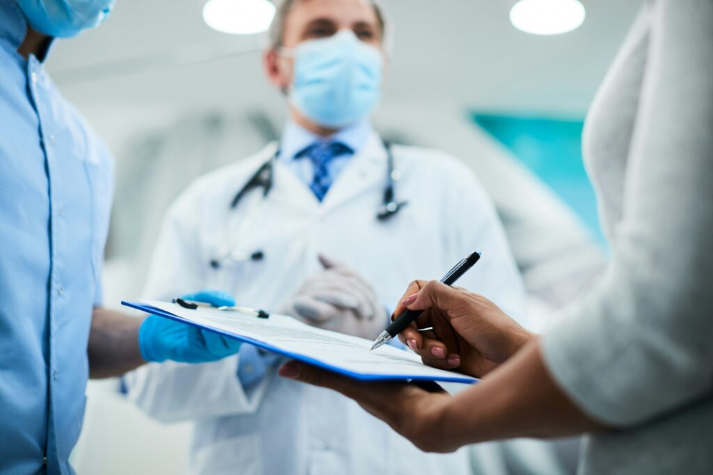 Close-up of a person holding a pen and signing a document on a clipboard held by a healthcare worker in a clinical setting, with another masked medical professional observing in the background.