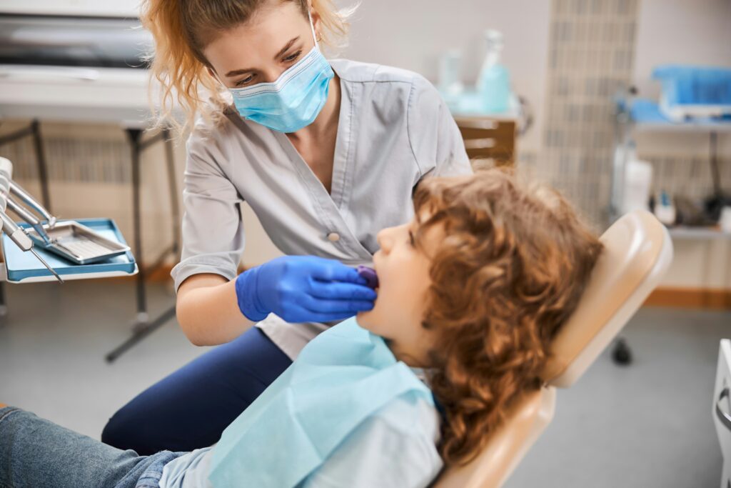 Female dentist wearing a face mask and gloves while examining a child patient in a dental clinic. The child sits in a dental chair during a routine check-up, with dental tools and equipment visible in the background.