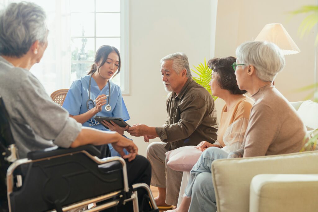 Healthcare worker speaking with an elderly family in a home care setting, using a tablet while discussing care and support, with warm natural lighting and a wheelchair visible in the foreground.