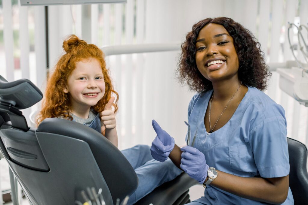 Smiling dentist and young patient giving thumbs up while sitting in a dental chair at a bright dental clinic, creating a friendly and positive treatment experience.