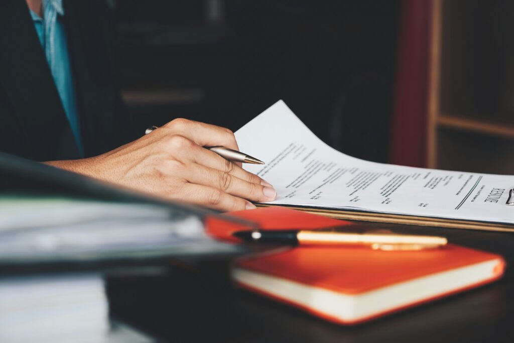 Close-up of a person reviewing and signing paperwork at a desk, holding a pen over a printed document with notebooks and office materials in the foreground.