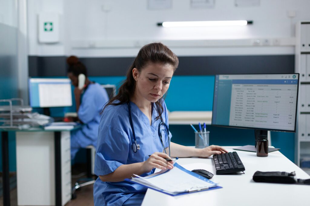 Nurse reviewing paperwork at a hospital workstation while using a computer, with another healthcare worker in the background.
