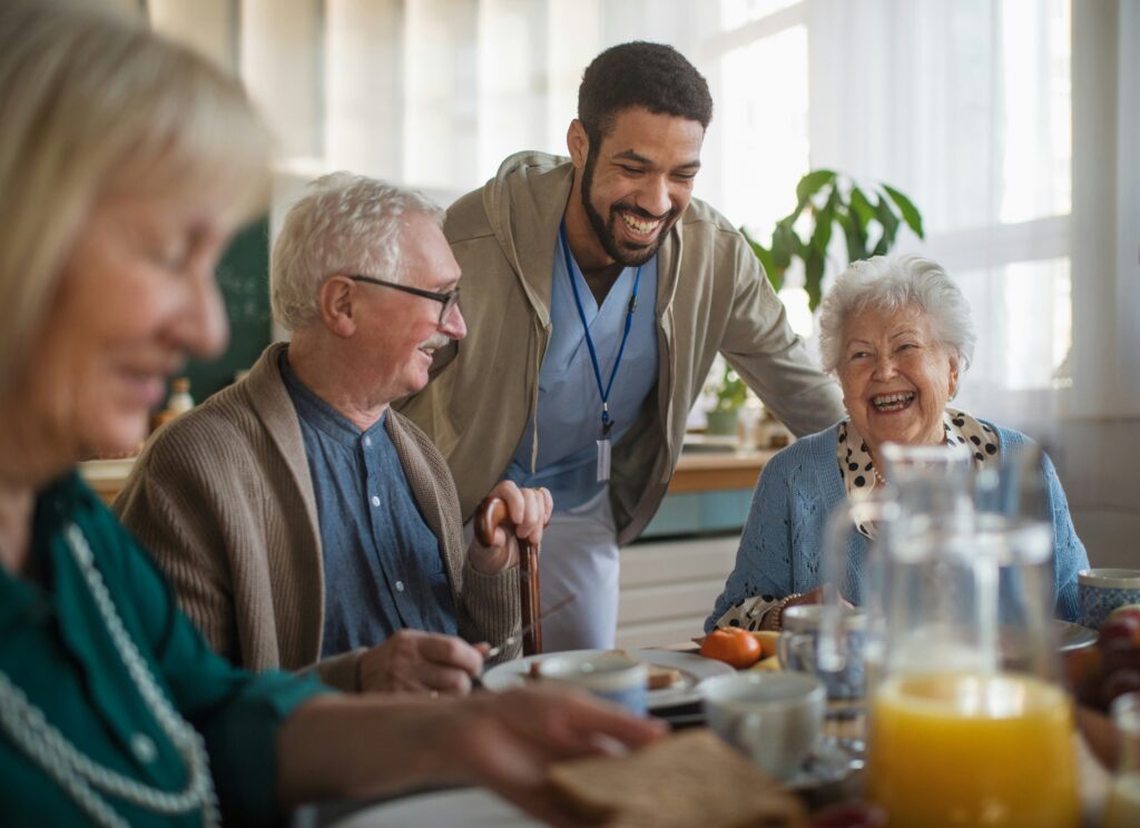 Healthcare worker smiling and interacting with elderly residents during a shared meal in an aged care setting, creating a warm and supportive atmosphere.
