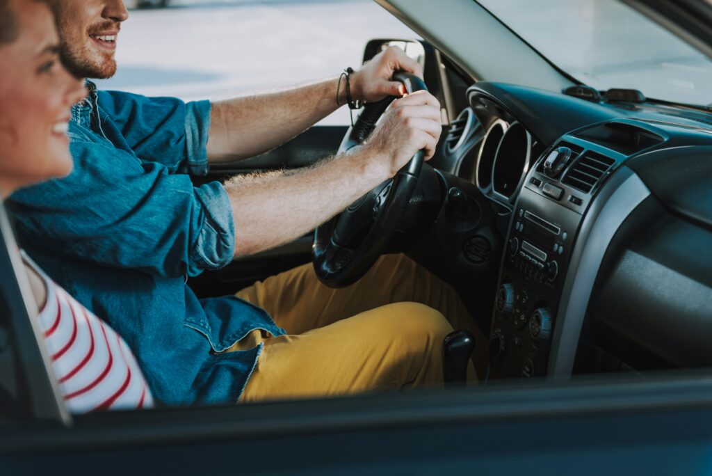 Close-up of a smiling man in a denim shirt driving a car, his hands on the steering wheel, with a passenger seated beside him.