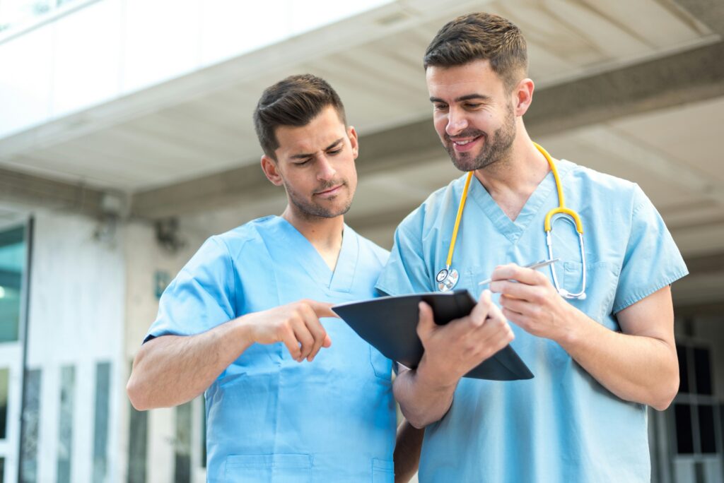 Two healthcare workers in blue scrubs reviewing information on a tablet outside a medical facility. One clinician points at the screen while the other holds a pen, with a stethoscope visible around their neck.