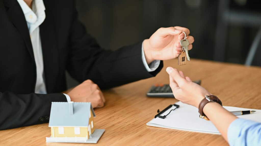 A mortgage broker handing house keys to a client across a desk, with a small model house, paperwork, and a calculator visible in the background.