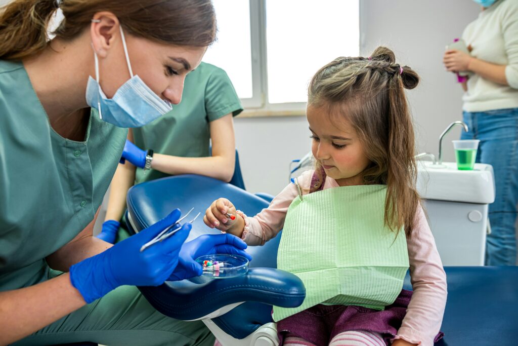 Female dental professional assisting a young child during a dental visit in a clinic, showing colourful dental tools while the child sits in a treatment chair wearing a protective bib.