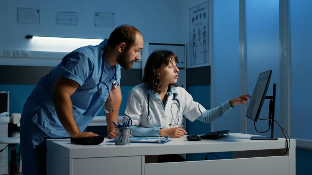 Two healthcare professionals reviewing information together at a computer during a late-night hospital shift, with medical equipment and soft blue clinical lighting in the background.