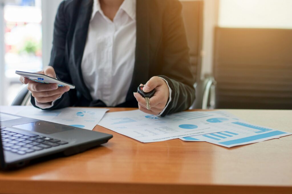 Business professional holding a car key and smartphone at a desk with financial documents and a laptop, representing car loan applications and finance planning.