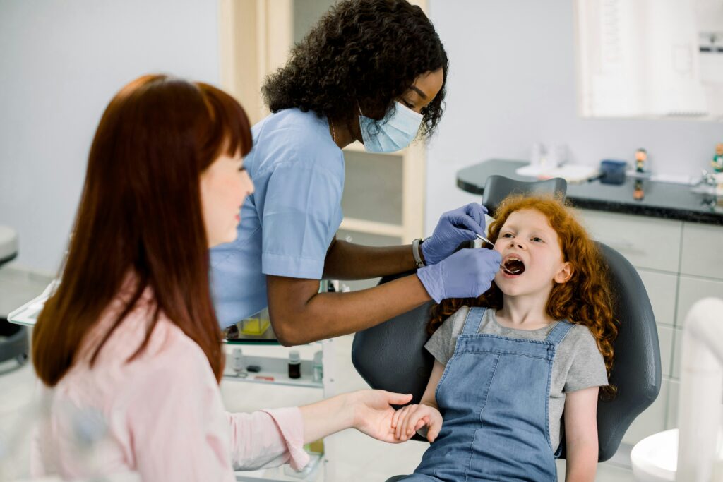 Dentist wearing a face mask and gloves while examining a young girl’s teeth in a dental clinic. The child sits in a dental chair holding her parent’s hand during the check-up.