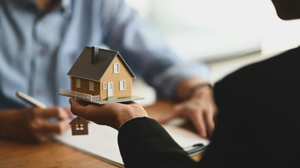 Close-up of a person holding a miniature house model during a home loan or property consultation, with paperwork and another person blurred in the background.