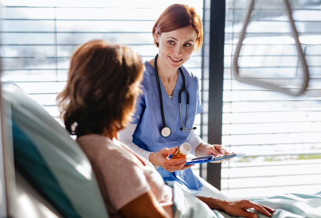 Nurse speaking with a patient in a hospital room while holding medication and reviewing notes on a clipboard.