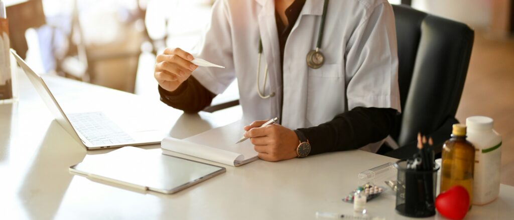 Doctor writing notes at a medical office desk with a laptop, tablet, and medication bottles nearby.
