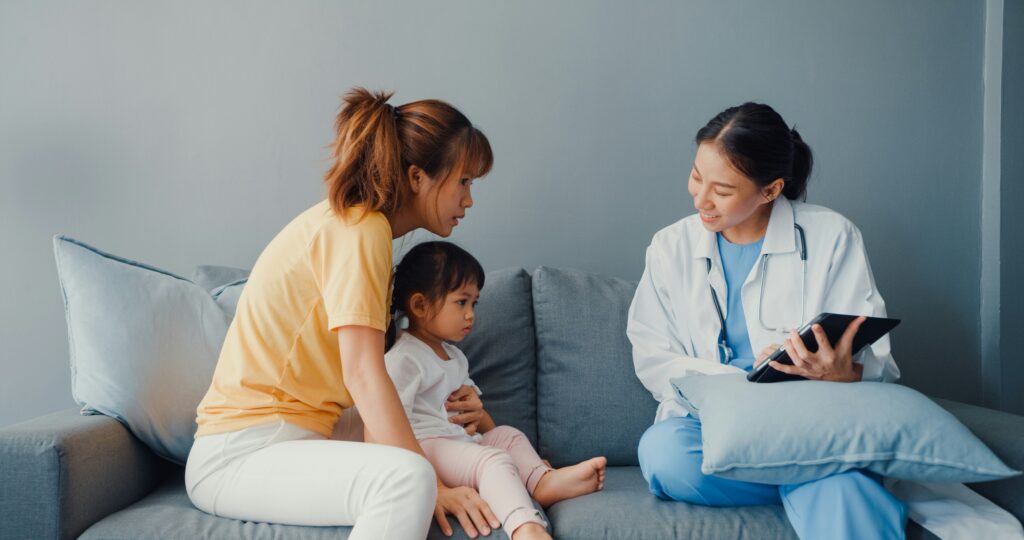 Female healthcare worker in a white coat speaking with a mother and young child during a home visit, holding a tablet while sitting on a couch in a living room.