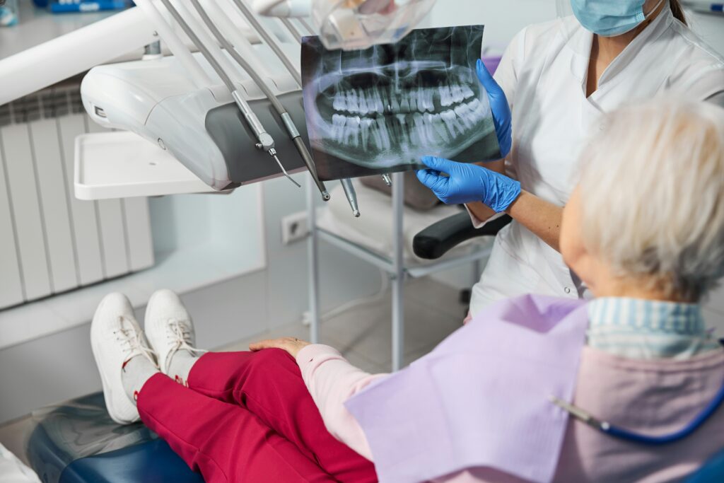 Dental professional wearing gloves and a face mask showing a dental X-ray scan to an elderly patient seated in a dental chair during a consultation in a modern clinic.