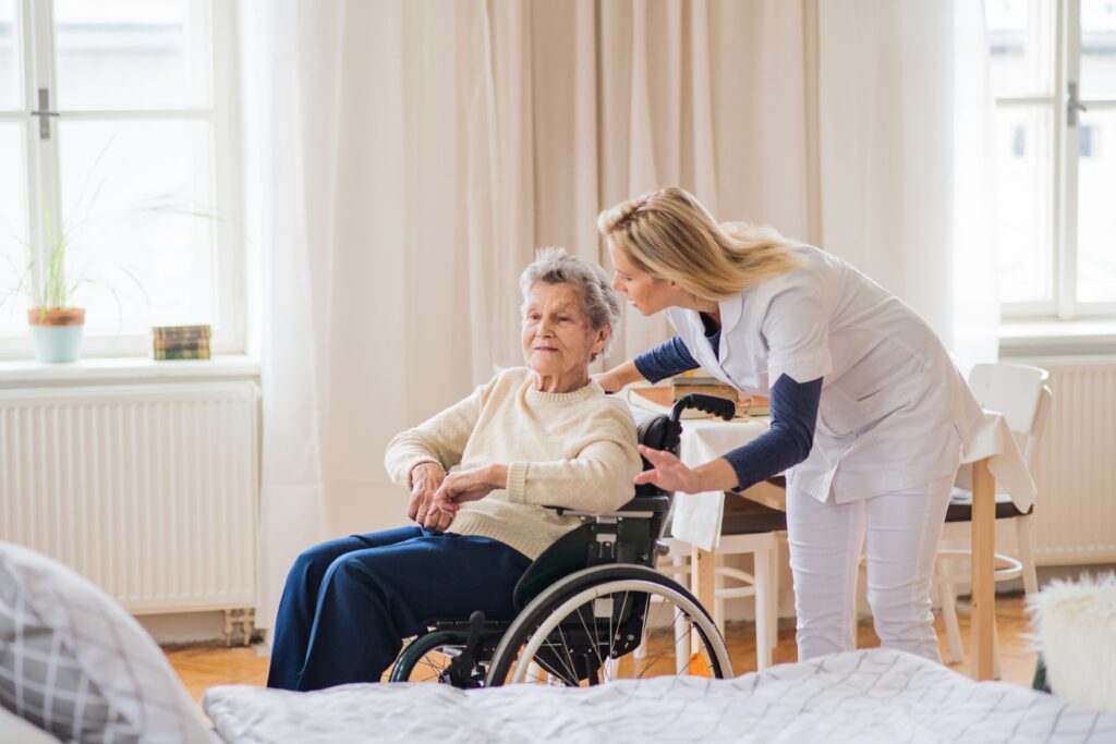 Healthcare worker assisting an elderly woman in a wheelchair inside a bright home setting, providing support and care beside a dining area.