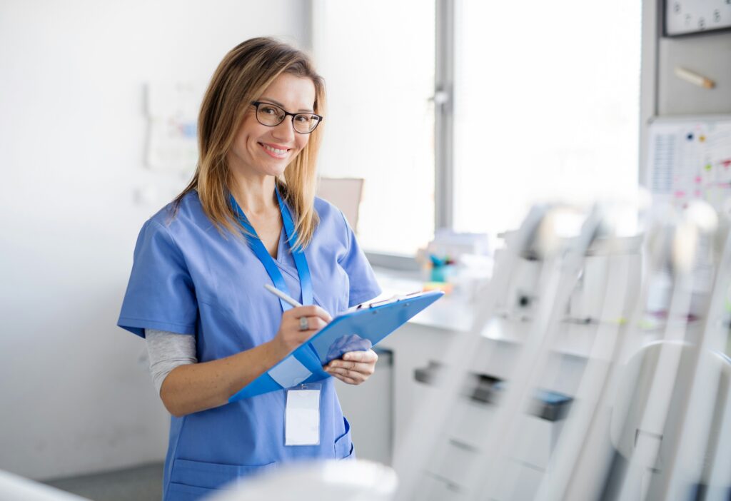 Smiling healthcare professional in blue scrubs holding a clipboard and pen in a bright clinic or medical office environment.