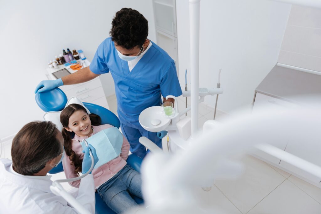 Dentist and dental assistant treating a smiling child patient in a clean dental clinic with modern equipment and bright lighting.