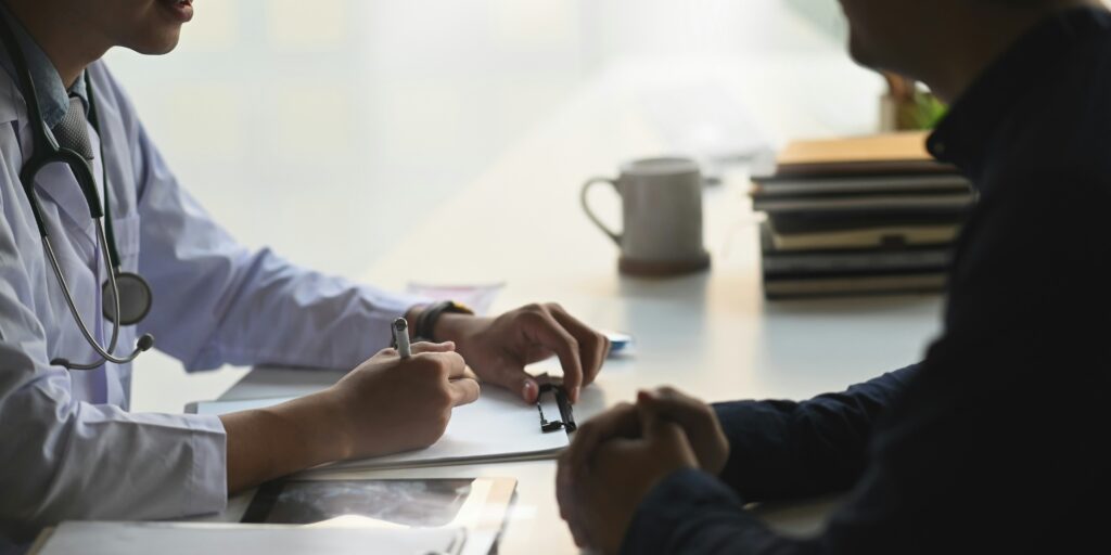 Doctor wearing a white coat and stethoscope speaking with a patient across a desk during a consultation, while writing notes on a clipboard in a softly lit medical office.
