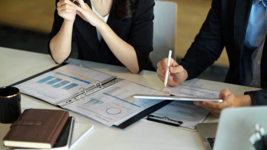 Two professionals reviewing financial documents and charts at a meeting table, with a tablet, clipboard, and reports spread across the desk in an office setting.