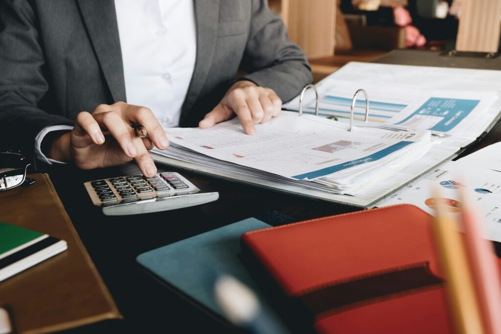 Business professional using a calculator while reviewing financial reports and charts at a desk with folders, documents, and office supplies. The image represents budgeting, accounting, or mortgage repayment planning in a professional office setting.