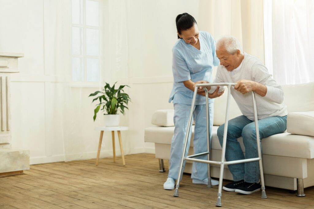 Healthcare worker in blue scrubs assisting an elderly man using a walking frame in a bright living room, helping him stand safely beside a couch.
