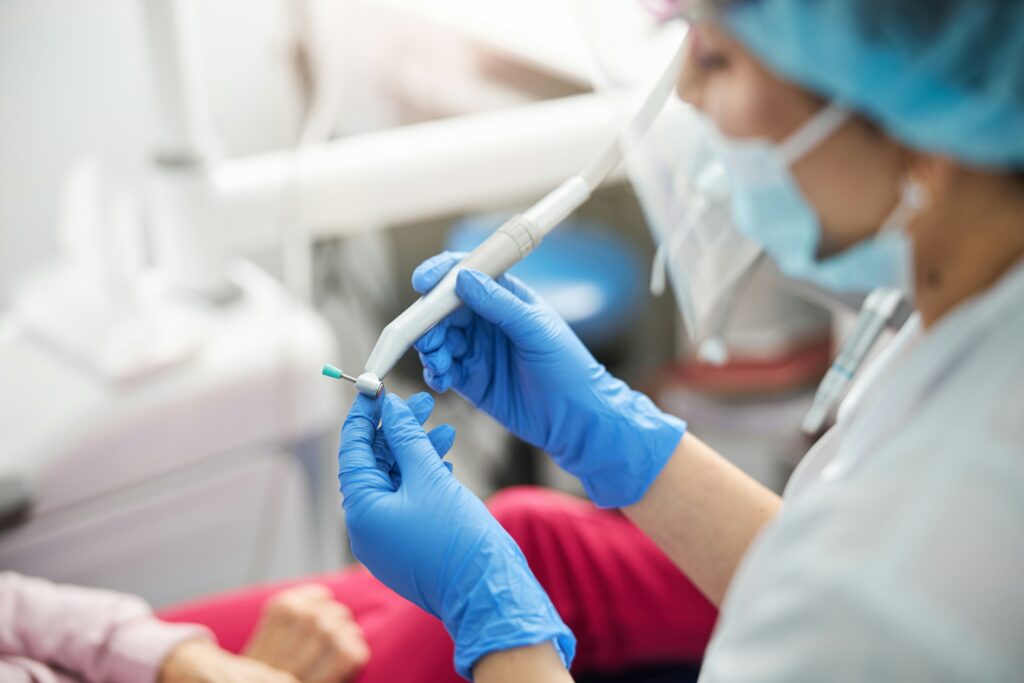 Close-up of a dental professional preparing a dental polishing tool while treating a patient in a clinical setting, with dental equipment visible in the background.