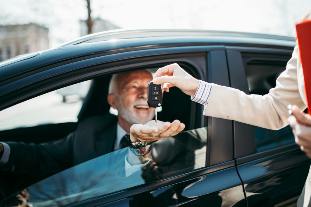 Close-up of a person handing car keys to an older man sitting inside a black vehicle, capturing the moment of receiving a new car. The driver is smiling through the open window while another person outside the car holds a clipboard, suggesting a car purchase, dealership handover, or vehicle delivery.