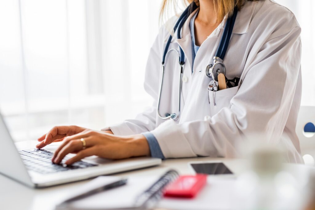 Doctor in a white coat typing on a laptop at a desk, with a stethoscope around their neck and office items including a phone and notebook nearby.