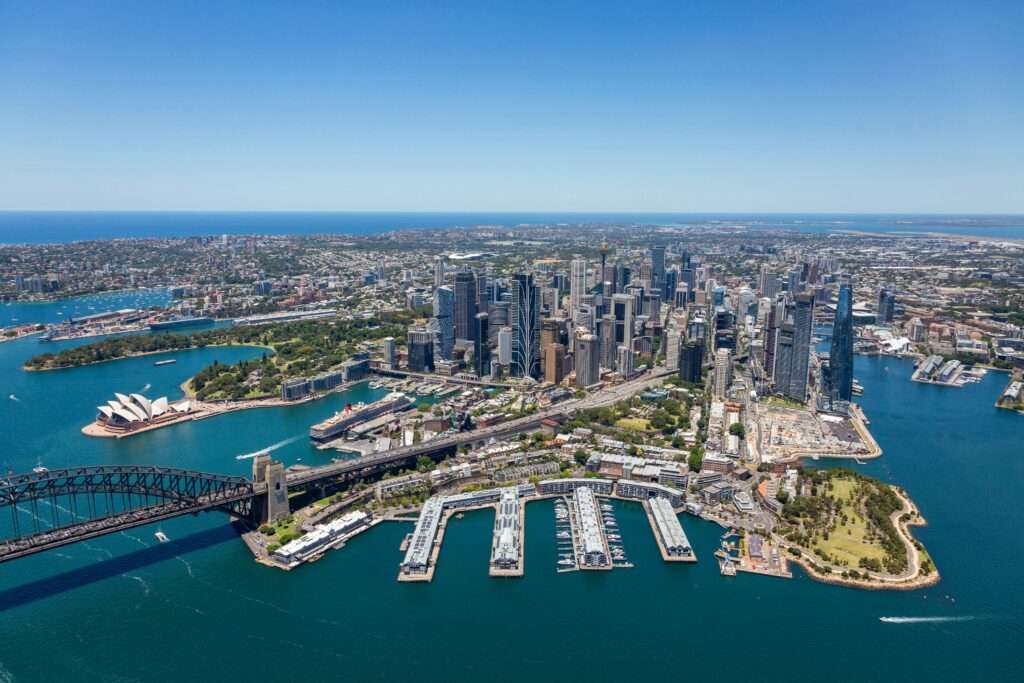 Aerial view of Sydney Harbour and Sydney CBD in New South Wales, Australia, featuring the Sydney Opera House, Harbour Bridge, waterfront skyline, and surrounding harbour on a clear sunny day.
