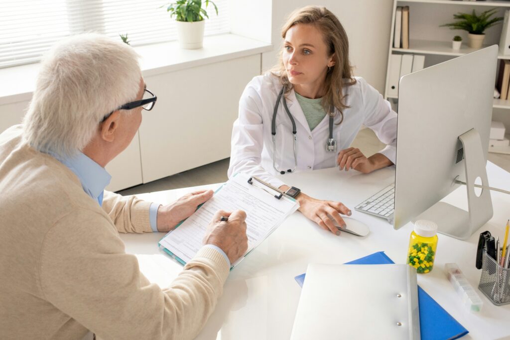 Doctor consulting with an elderly patient in a medical office while reviewing paperwork at a computer desk.