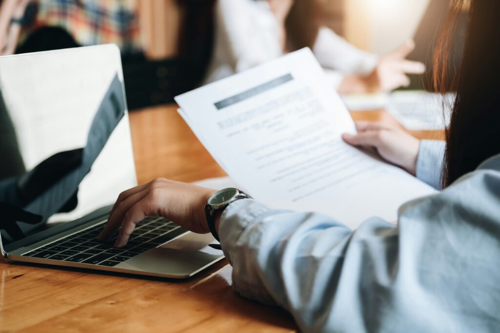 Person reviewing printed financial documents while working on a laptop at a wooden desk during a business meeting.