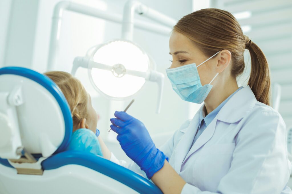 Female dentist wearing a face mask and gloves while treating a child patient in a modern dental clinic. The child sits in a dental chair as the dentist prepares dental tools under an examination light.