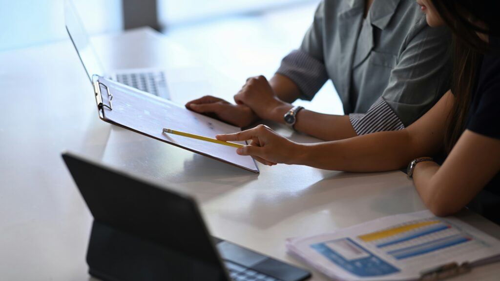 Two people sitting at a desk reviewing financial documents on a clipboard beside open laptops and paperwork in a modern office setting.