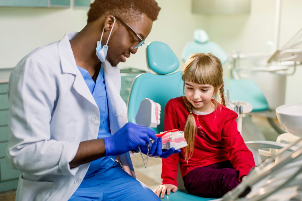 Dentist in a dental clinic demonstrating a large model of teeth and a toothbrush to a young girl seated in a dental chair during a children’s dental consultation.