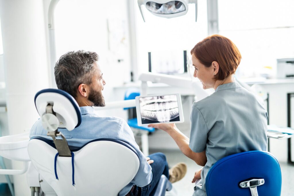 Dentist discussing dental X-ray results with a male patient seated in a modern dental clinic, using a digital screen to explain the patient’s oral health condition.