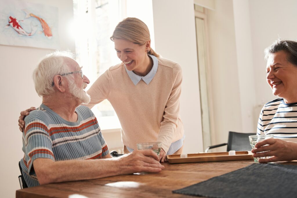 Healthcare worker warmly interacting with elderly residents around a dining table in a home care or aged care environment, creating a supportive and compassionate atmosphere.