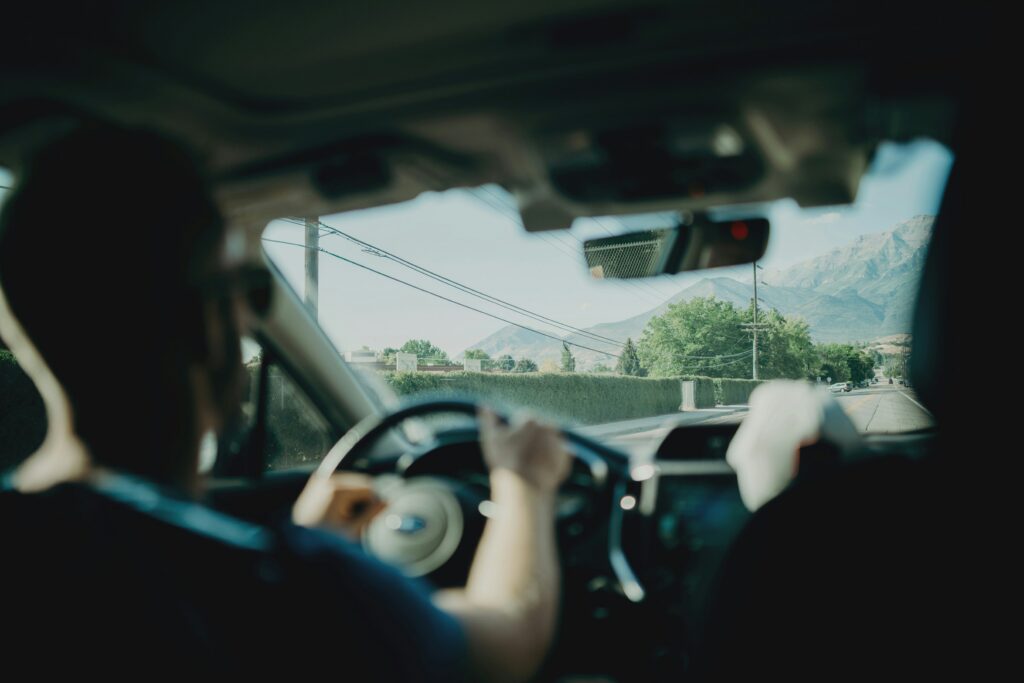 View from inside a moving car with a driver holding the steering wheel, looking out toward a quiet suburban road and distant mountains through the windshield.