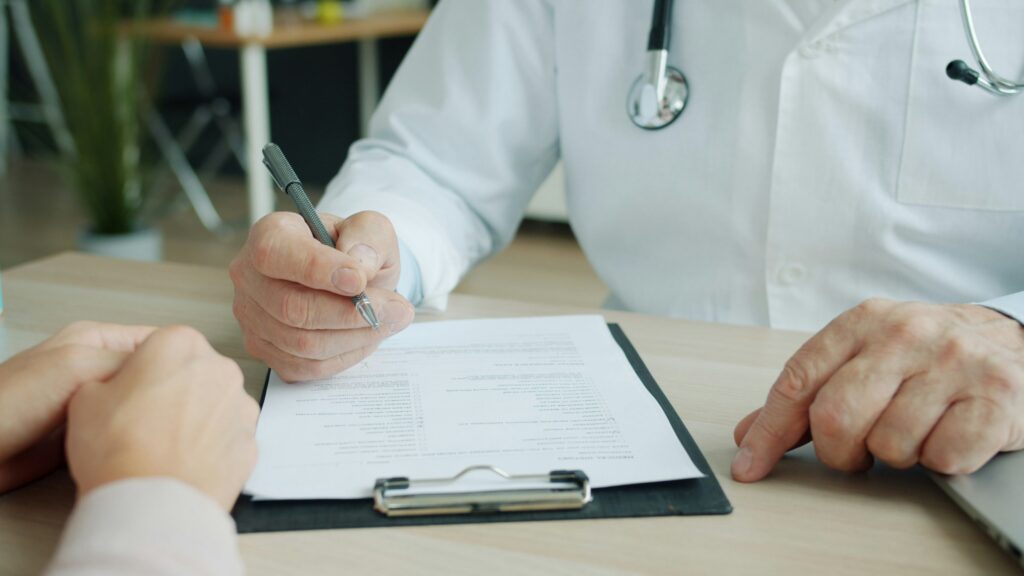 Doctor consulting with a patient while reviewing paperwork on a clipboard in a medical office. The healthcare professional points to a document during a discussion, with a stethoscope visible on their white coat.